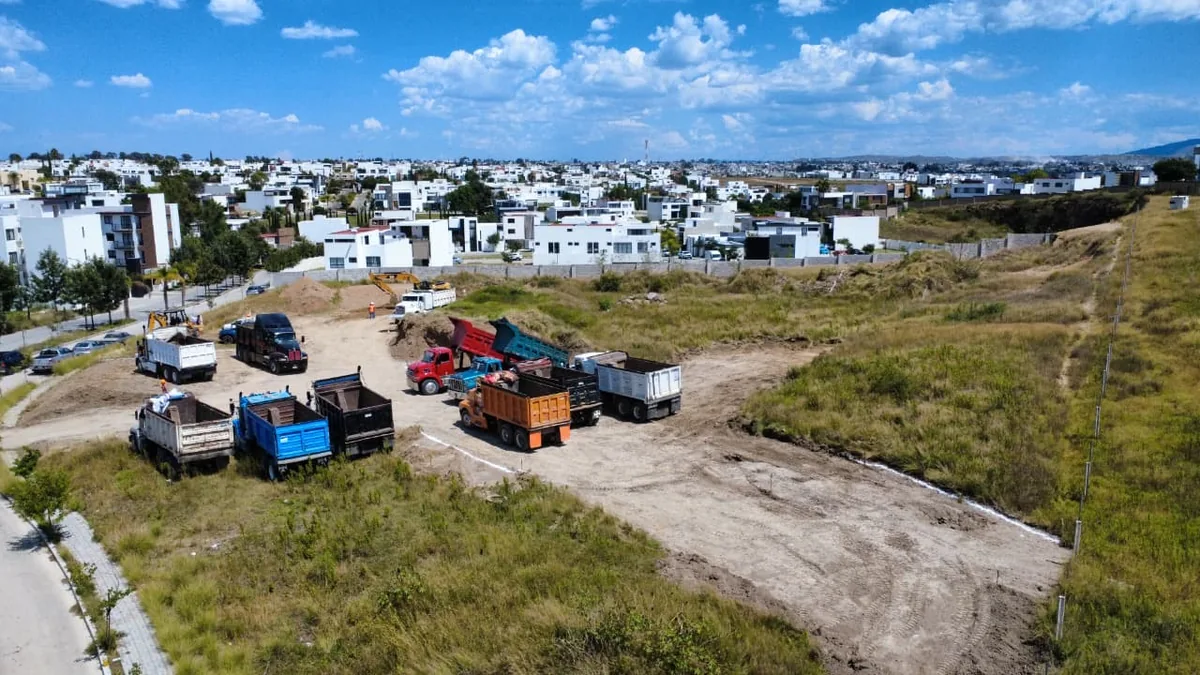 Camiones de volteo y retroexcavadoras en el terreno, con viviendas de la zona residencial al fondo