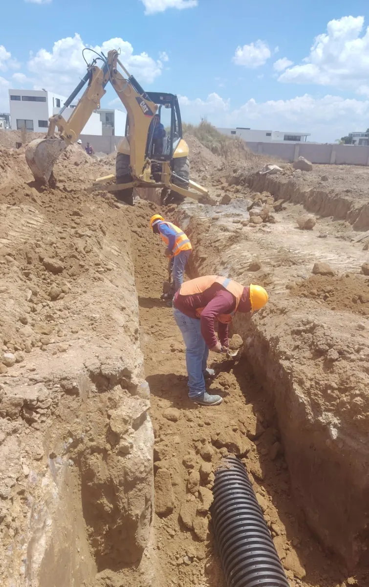 Retroexcavadora abriendo zanja mientras dos trabajadores afinan con pala; tubería corrugada en primer plano